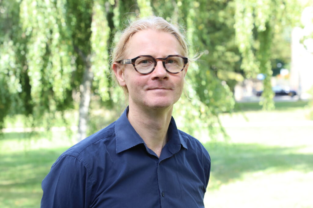 blond man with brown glasses wearing a blue shirt , blurry green background - portrait