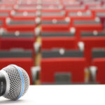 picture of an empty lecture hall with red seats blurry in the background and a microphone lying on the desk in the front