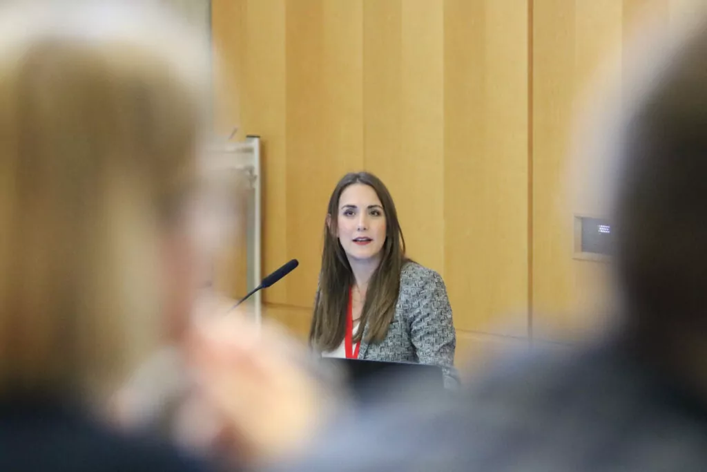 Woman with long brown hair standing at a desk in the lecture hall giving a presentation