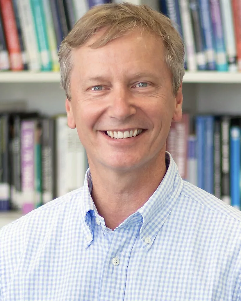 blond man in front of a book shelf wearing a light blue checkered shirt