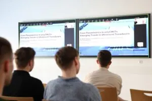 4 männliche Personen schauen auf zwei Bildschirme während eines Vortrages.